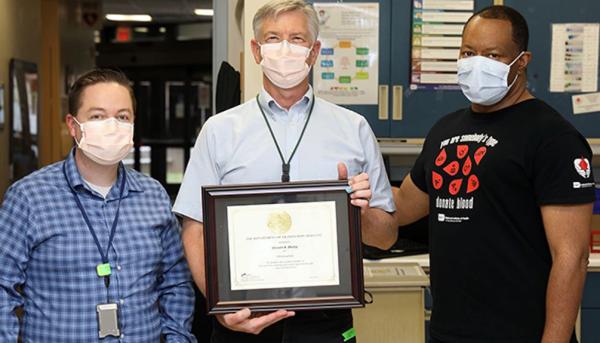 Steven Bailey posing with DTM clinical fellow Dr. James Long (left) and donor recruitment supervisor Hal Wilkens (right) as he receives a plaque commemorating his 100th blood donation at the NIH Blood Bank