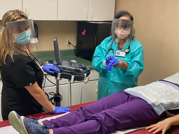 Two health care workers wearing cloth masks and face shields with a patient who is lying down and whose face is not visible