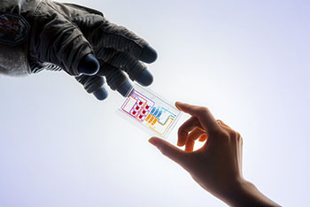 a translucent rectangular plate holding a tissue chip; the chip is being handed from one hand to another wearing an astronaut glove