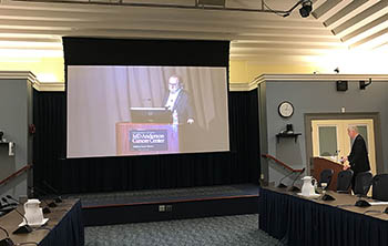 movie screen showing James Allison at a podium; NIH Director Francis Collins is watching from Wilson Hall at NIH