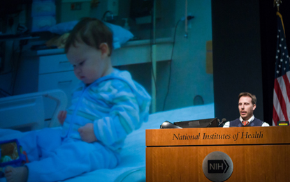  MATT MIGHT AT PODIUM; IMAGE OF CHILD IN A HOSPITAL ON SCREEN BEHIND MIGHT