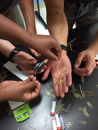 Several sets of hands working with a battery and paper clips to make an electromagnet