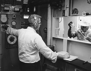 man with headphones observing scientist in anaerobic lab through a window